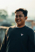 A smiling person wearing a Gratitude branded sweatshirt, standing in a sunlit park.