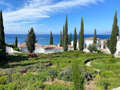 A coastal scene featuring several white buildings with terracotta roofs surrounded by tall, slender cypress trees. The foreground displays a well-manicured garden with vibrant green shrubs and pathways. In the background, a vast expanse of blue ocean stretches towards the horizon, topped with a partly cloudy sky.