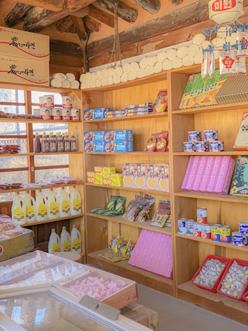 A traditional market stall with wooden shelves is stocked with a variety of packaged goods, including snacks, canned foods, and bottles of liquids. The shelves display colorful products, some with Korean text, and the ceiling shows wooden beams. The overall setting is bright and filled with neatly arranged items, giving a sense of an organized and well-stocked market.