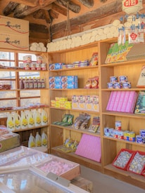 A traditional market stall with wooden shelves is stocked with a variety of packaged goods, including snacks, canned foods, and bottles of liquids. The shelves display colorful products, some with Korean text, and the ceiling shows wooden beams. The overall setting is bright and filled with neatly arranged items, giving a sense of an organized and well-stocked market.