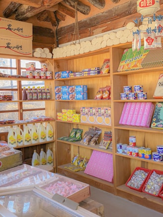 A traditional market stall with wooden shelves is stocked with a variety of packaged goods, including snacks, canned foods, and bottles of liquids. The shelves display colorful products, some with Korean text, and the ceiling shows wooden beams. The overall setting is bright and filled with neatly arranged items, giving a sense of an organized and well-stocked market.