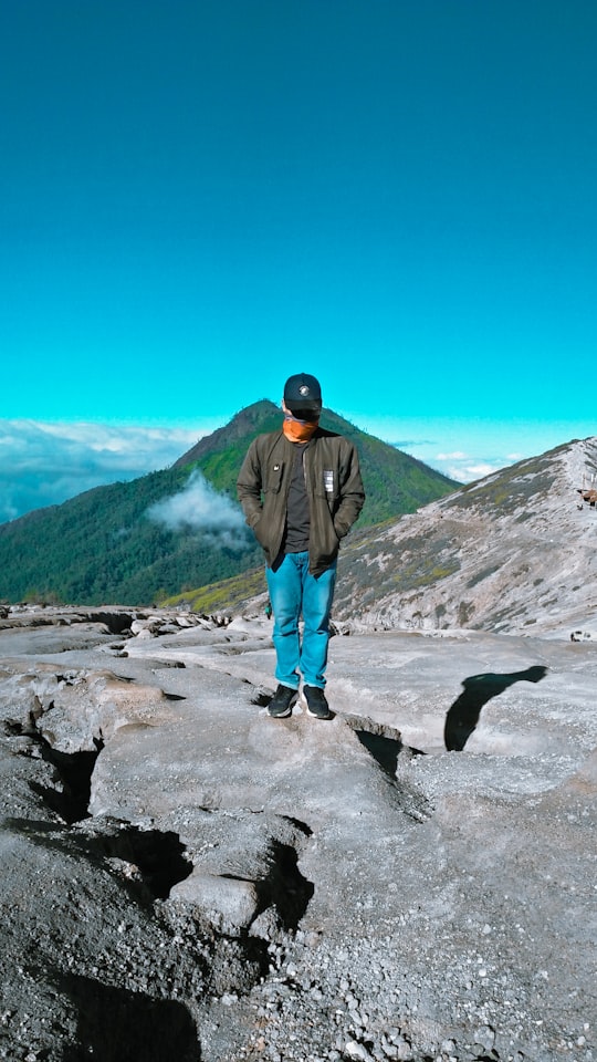 man in black jacket and blue denim jeans standing on rocky mountain during daytime in Ijen Indonesia