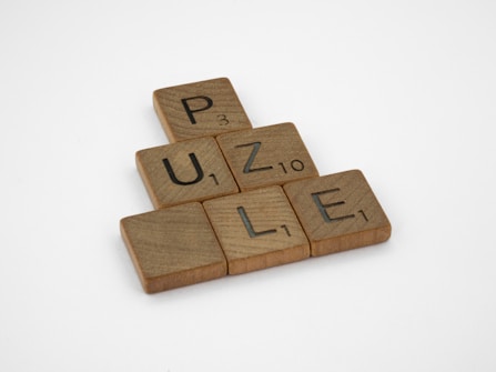 Wooden Scrabble tiles spelling out the word 'PUZZLE' are arranged in a triangular formation on a plain white background. Each tile has a letter with a number indicating its point value in the game.
