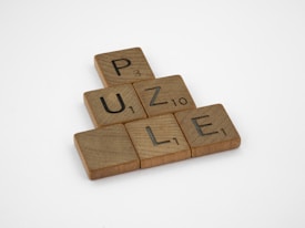 Wooden Scrabble tiles spelling out the word 'PUZZLE' are arranged in a triangular formation on a plain white background. Each tile has a letter with a number indicating its point value in the game.