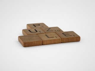 Wooden letter tiles spell out the word 'PUZZLE' on a flat surface. Each tile has a different letter with a small number in the corner, resembling Scrabble game pieces.