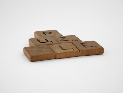 Wooden letter tiles spell out the word 'PUZZLE' on a flat surface. Each tile has a different letter with a small number in the corner, resembling Scrabble game pieces.
