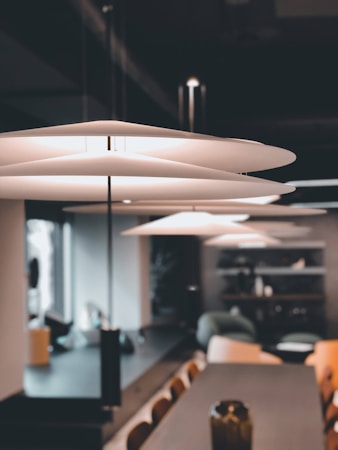 A modern interior space featuring a sleek, contemporary pendant light with layered, disc-shaped shades. The setting includes a long wooden table surrounded by chairs, a vase in the foreground, and a comfortable seating area with shelves in the background. Natural light streams in through a window, creating a warm and inviting atmosphere.