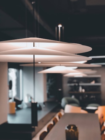 A modern interior space featuring a sleek, contemporary pendant light with layered, disc-shaped shades. The setting includes a long wooden table surrounded by chairs, a vase in the foreground, and a comfortable seating area with shelves in the background. Natural light streams in through a window, creating a warm and inviting atmosphere.
