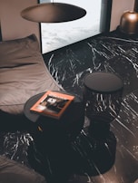 A modern interior with a black marble floor featuring white veins. A dark round table holds a book titled 'New Romance' with an orange cover. A large dark brown cushion or beanbag lies next to the table, under a minimalist ceiling light fixture. A bronze vase is positioned in the corner, adding a metallic accent to the sophisticated decor.