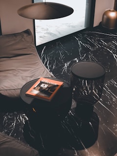 A modern interior with a black marble floor featuring white veins. A dark round table holds a book titled 'New Romance' with an orange cover. A large dark brown cushion or beanbag lies next to the table, under a minimalist ceiling light fixture. A bronze vase is positioned in the corner, adding a metallic accent to the sophisticated decor.