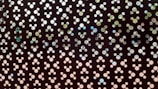 A perforated metal sheet with a repeating pattern of circular holes in various sizes. Through the holes, glimpses of a natural setting with hints of trees and sky are visible. The arrangement creates an abstract and artistic design.