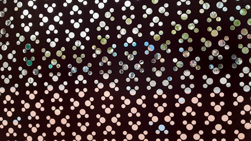 A perforated metal sheet with a repeating pattern of circular holes in various sizes. Through the holes, glimpses of a natural setting with hints of trees and sky are visible. The arrangement creates an abstract and artistic design.