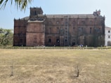A large, historic brick building with intricate architectural details and multiple windows. It features a prominent central tower and is surrounded by a grass courtyard and palm trees. There is scaffolding on one side, indicating maintenance or restoration work.