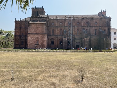 A large, historic brick building with intricate architectural details and multiple windows. It features a prominent central tower and is surrounded by a grass courtyard and palm trees. There is scaffolding on one side, indicating maintenance or restoration work.