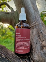 A brown glass bottle labeled 'kiara Pain Relief' is placed on a tree branch. The bottle has a white screw cap with a dropper. The label describes the contents as a full spectrum tincture, listing ingredients such as CBD and THC, along with usage instructions.