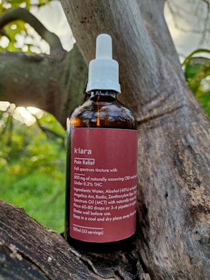 A brown glass bottle labeled 'kiara Pain Relief' is placed on a tree branch. The bottle has a white screw cap with a dropper. The label describes the contents as a full spectrum tincture, listing ingredients such as CBD and THC, along with usage instructions.