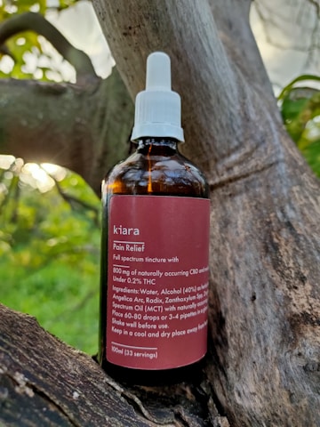 A brown glass bottle labeled 'kiara Pain Relief' is placed on a tree branch. The bottle has a white screw cap with a dropper. The label describes the contents as a full spectrum tincture, listing ingredients such as CBD and THC, along with usage instructions.