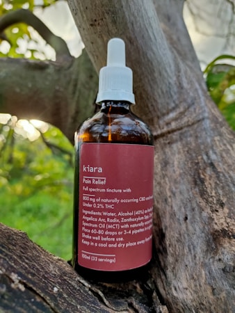 A brown glass bottle labeled 'kiara Pain Relief' is placed on a tree branch. The bottle has a white screw cap with a dropper. The label describes the contents as a full spectrum tincture, listing ingredients such as CBD and THC, along with usage instructions.