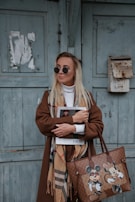 A stylish woman wearing sunglasses and a long brown coat stands against a weathered green wooden door. She holds a magazine with a Dior cover and carries a designer handbag featuring cartoon characters. Her scarf adds a touch of elegance with a classic pattern.