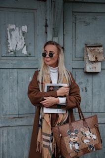 A stylish woman wearing sunglasses and a long brown coat stands against a weathered green wooden door. She holds a magazine with a Dior cover and carries a designer handbag featuring cartoon characters. Her scarf adds a touch of elegance with a classic pattern.