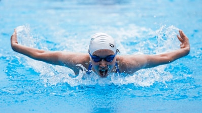 A swimmer is performing the butterfly stroke in a swimming pool, with water splashing around them as their arms are outstretched and their face is partially submerged. They are wearing a white swim cap and blue goggles.