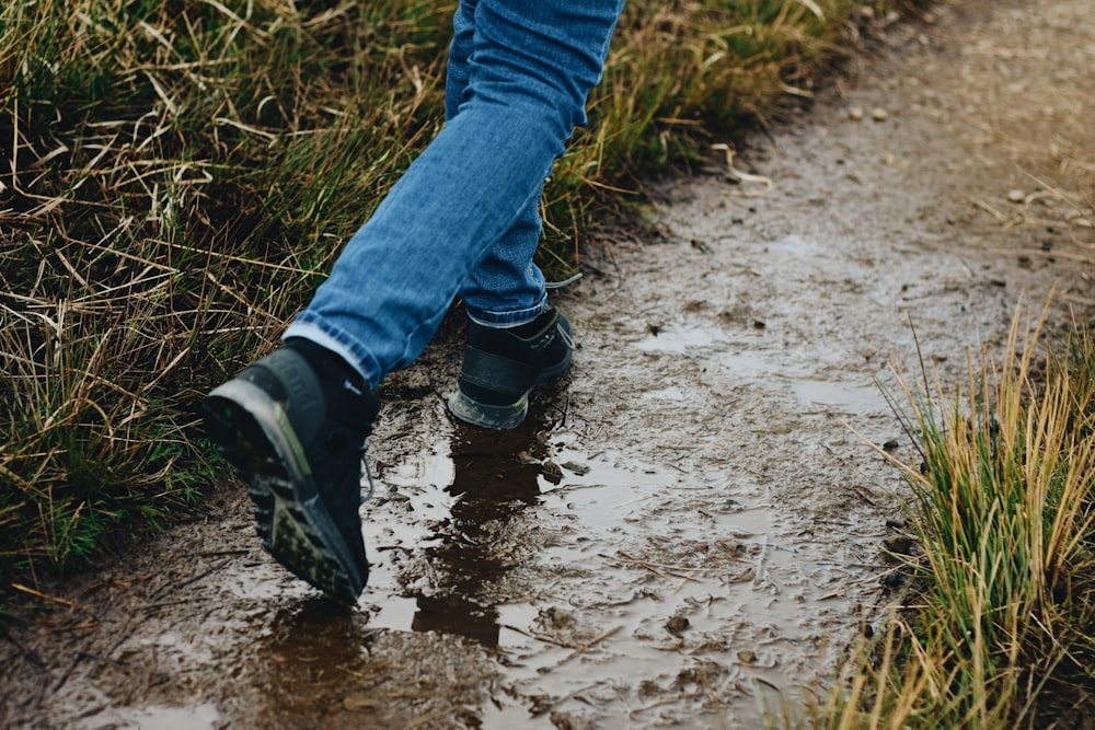 person in blue denim jeans and black leather boots walking on wet dirt road