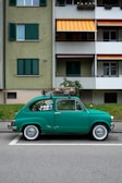 A vintage green car is parked on a city street in front of an apartment building. The car is covered with various stickers on the rear side window, and there is luggage strapped to the roof. The building behind has green and white exterior walls with closed shutters and colorful striped awnings.