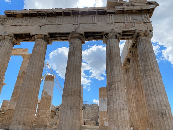Ancient stone columns with a classical architectural style rise majestically against a backdrop of a bright blue sky with scattered clouds. A construction crane is visible, indicating ongoing restoration work. The columns are weathered, displaying a sense of historical grandeur and timeless beauty.