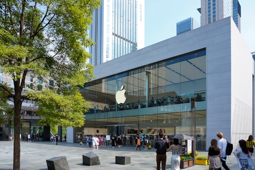 A modern glass-fronted store with a prominent logo on the facade, bustling with people. It features a large tree in the foreground and is set against a backdrop of tall city buildings.