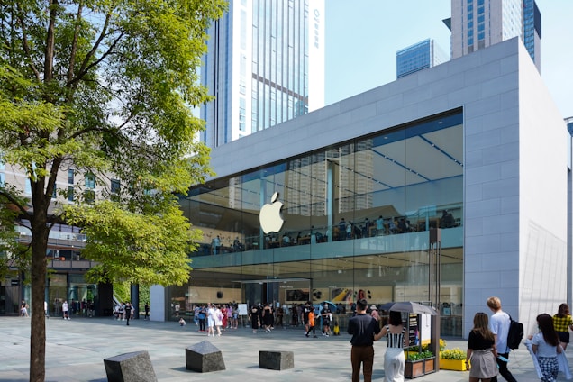 A modern glass-fronted store with a prominent logo on the facade, bustling with people. It features a large tree in the foreground and is set against a backdrop of tall city buildings.