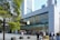 A modern glass-fronted store with a prominent logo on the facade, bustling with people. It features a large tree in the foreground and is set against a backdrop of tall city buildings.
