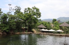 A tranquil outdoor setting featuring a natural pool surrounded by lush greenery and trees. Thatched umbrellas are positioned near the water's edge, and mountains can be seen in the background under a partly cloudy sky.