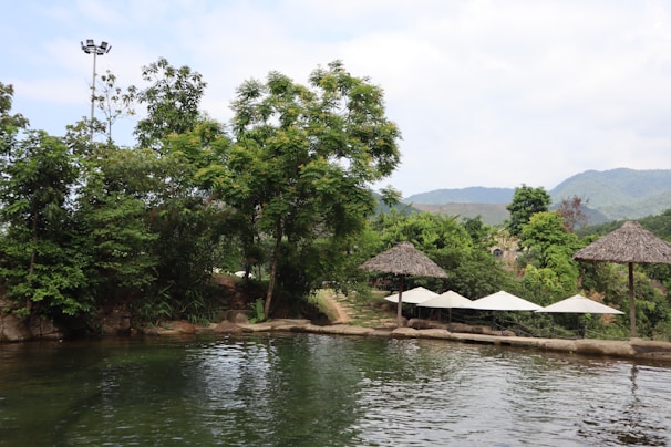 A tranquil outdoor setting featuring a natural pool surrounded by lush greenery and trees. Thatched umbrellas are positioned near the water's edge, and mountains can be seen in the background under a partly cloudy sky.