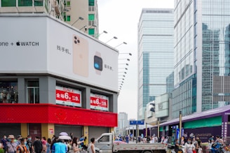 An urban street scene features a large building with a prominent advertisement for smartphones and smartwatches on the corner. Below the advertisement, a red horizontal strip displays sale signs. The street is bustling with people, many of whom are wearing masks. In the background, there are several high-rise buildings with glass facades.
