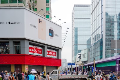 An urban street scene features a large building with a prominent advertisement for smartphones and smartwatches on the corner. Below the advertisement, a red horizontal strip displays sale signs. The street is bustling with people, many of whom are wearing masks. In the background, there are several high-rise buildings with glass facades.