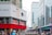 An urban street scene features a large building with a prominent advertisement for smartphones and smartwatches on the corner. Below the advertisement, a red horizontal strip displays sale signs. The street is bustling with people, many of whom are wearing masks. In the background, there are several high-rise buildings with glass facades.
