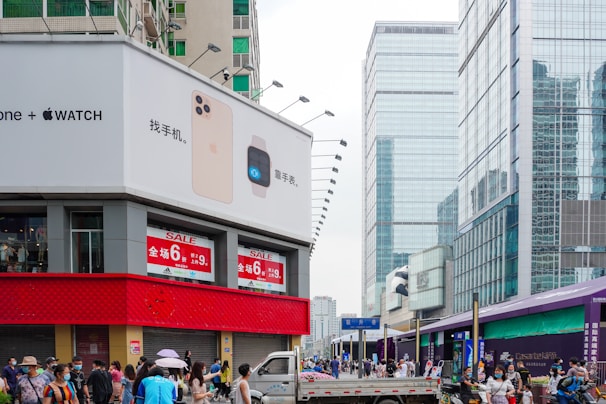 An urban street scene features a large building with a prominent advertisement for smartphones and smartwatches on the corner. Below the advertisement, a red horizontal strip displays sale signs. The street is bustling with people, many of whom are wearing masks. In the background, there are several high-rise buildings with glass facades.