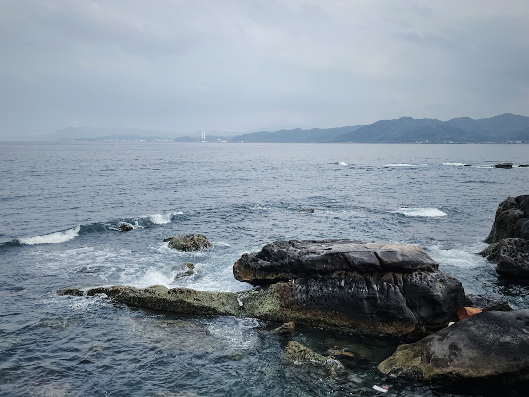 新北市貢寮區東北角海岸的黑色岩礁與浪花，遠方是連綿的山線