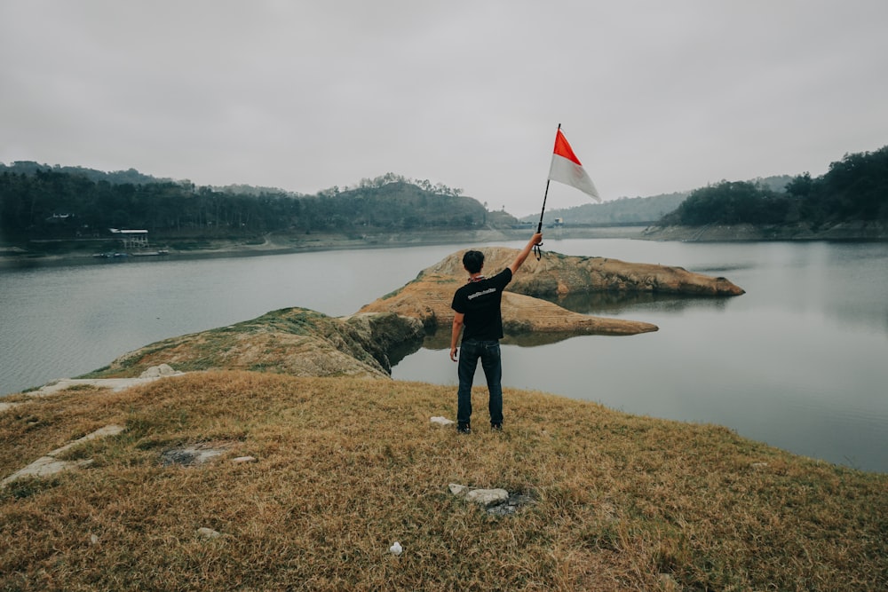 woman in black jacket and black pants holding flag standing on rock near lake during daytime