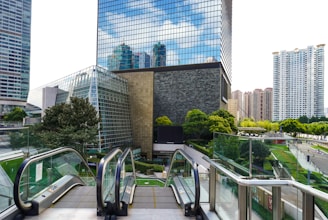 A modern urban scene featuring tall glass skyscrapers with reflective surfaces surrounded by other tall buildings. There are green trees and landscaped open spaces, with an escalator leading downward in the foreground.