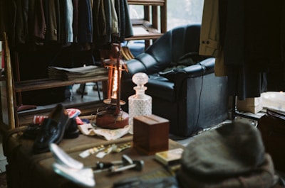 A cozy room filled with various items, including a rack of hanging clothes, a leather armchair, a vintage lamp, newspapers, and a crystal decanter on a wooden table. The light creates a warm atmosphere, highlighting the eclectic mix of household and personal items, such as shoes, hats, and small tools.