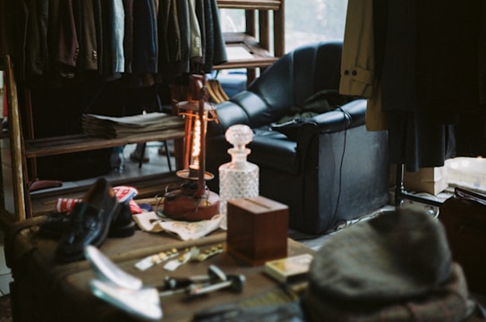 A cozy room filled with various items, including a rack of hanging clothes, a leather armchair, a vintage lamp, newspapers, and a crystal decanter on a wooden table. The light creates a warm atmosphere, highlighting the eclectic mix of household and personal items, such as shoes, hats, and small tools.