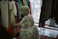 A person with blonde hair is looking through a rack of colorful dresses in a well-lit room. The dresses vary in patterns and colors, including florals, stripes, and solids. The individual is wearing a floral dress and appears to be in a vintage or thrift store setting. The scene is cozy and has a nostalgic atmosphere.