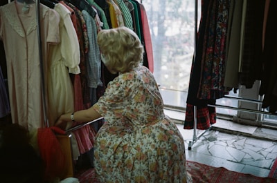 A person with blonde hair is looking through a rack of colorful dresses in a well-lit room. The dresses vary in patterns and colors, including florals, stripes, and solids. The individual is wearing a floral dress and appears to be in a vintage or thrift store setting. The scene is cozy and has a nostalgic atmosphere.