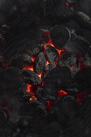 A close-up view of glowing charcoal briquettes with a warm, orange-red glow emanating from between the blackened coals. The texture of the charcoal is rough and ashy, highlighting the contrast between the dark surface and the vibrant heat.