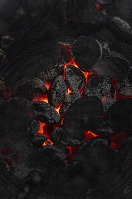 A close-up view of glowing charcoal briquettes with a warm, orange-red glow emanating from between the blackened coals. The texture of the charcoal is rough and ashy, highlighting the contrast between the dark surface and the vibrant heat.