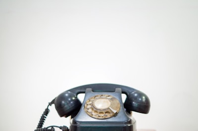 A vintage rotary dial telephone with a black body and earpiece is centered against a plain light background. The phone's dial, which has a beige tint, is prominently displayed at the top.