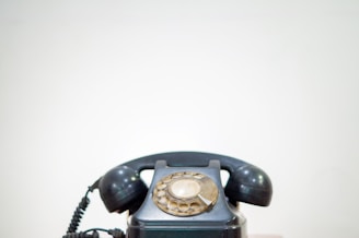 A vintage rotary dial telephone with a black body and earpiece is centered against a plain light background. The phone's dial, which has a beige tint, is prominently displayed at the top.