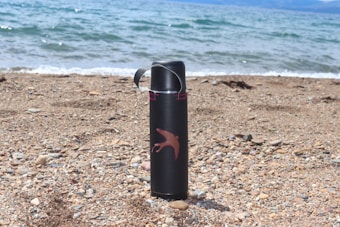 A black thermos with a red bird design rests upright on a pebbly beach with gentle waves meeting the shore in the background.