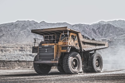 A large yellow dump truck is situated in an expansive quarry or mining site, surrounded by rocky, mountainous terrain. The truck is stationary, with dust or smoke around it, indicating recent movement.
